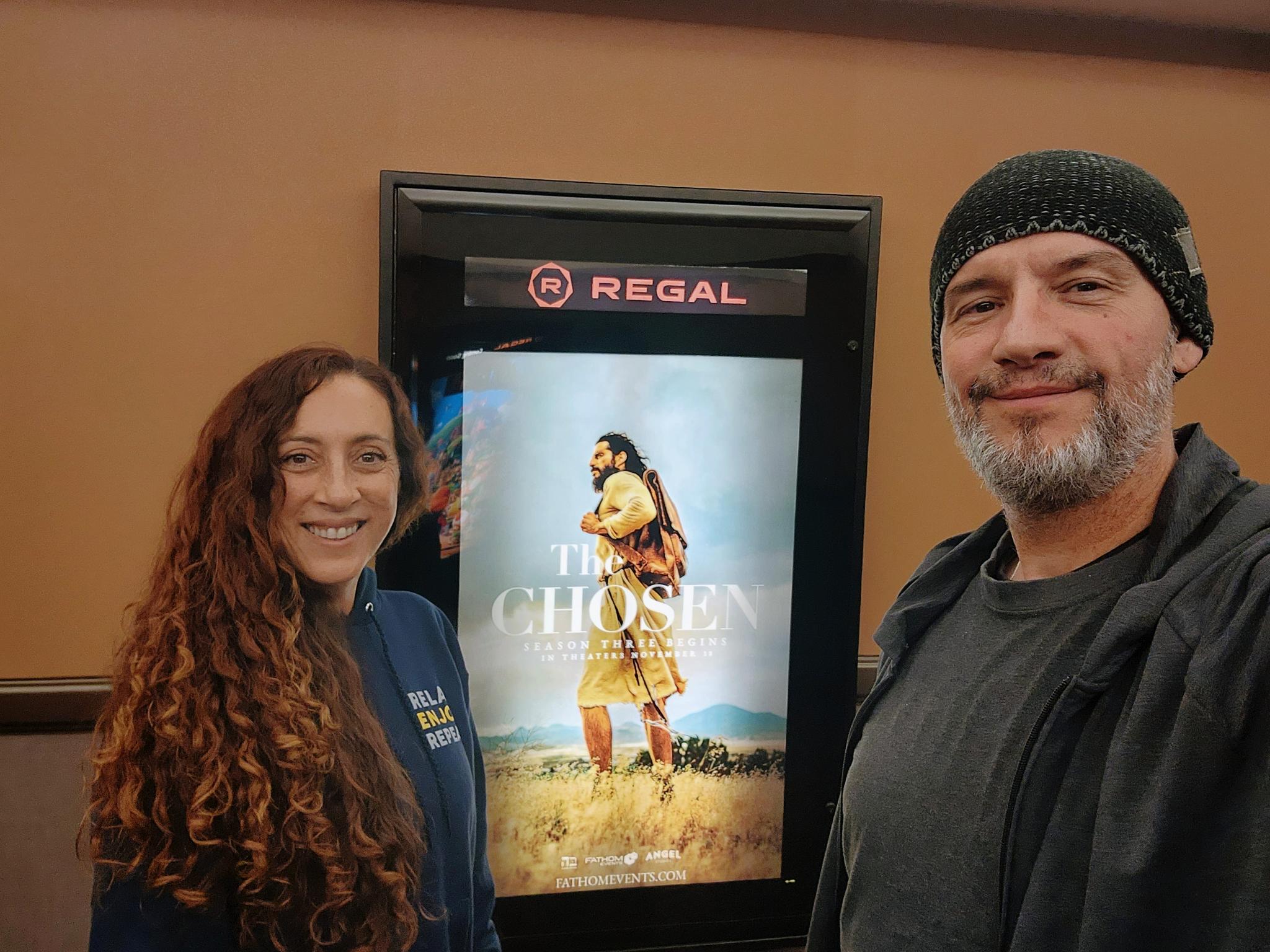 Andie and Greg beside a poster for The Chosen